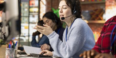 Call center operator next to coworkers dealing with upset customers, speaking using headset at office desk. Teamworking staff members doing customer support job, handling clients complaints