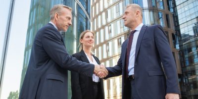 Confident middle-aged businessmen greeting each other and handshaking. Successful content young businesswoman standing near them and smiling. Building on background. Teamwork and partnership concept