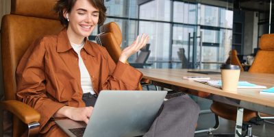 Portrait of woman discussing work project during online meeting, looking at laptop, working in office, gesturing while talking to coworker.